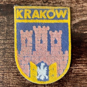 May include: A rectangular fabric patch with rounded bottom corners featuring the text KRAKOW in yellow letters on a blue background. The center displays a pinkish-tan castle illustration with three towers above a white eagle emblem on blue.