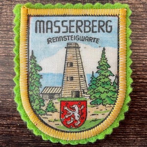 Può includere: Una toppa in tessuto rettangolare con bordo in feltro verde lime smerlato e rifinitura ricamata gialla. Il centro presenta un'illustrazione stampata di una torre, alberi di pino e uno scudo rosso con un leone bianco, etichettata MASSERBERG RENNSTEIGWARTE.