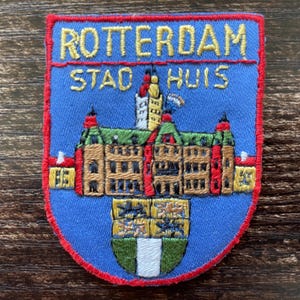 Peut inclure: Un écusson brodé en forme de bouclier avec un fond bleu et une bordure rouge. Il présente le texte jaune ROTTERDAM STAD HUIS au-dessus d'une représentation d'un bâtiment au toit vert et d'un blason en bas avec des lions et une bande blanche.