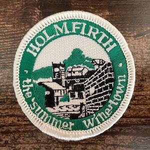 May include: A circular embroidered patch featuring a green border with white text that reads HOLMFIRTH and the summer wine town. The centre depicts a black and white stitched illustration of a stone building with a tower and a brick wall.
