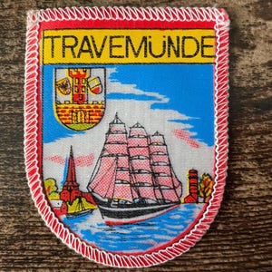 May include: A rectangular embroidered patch with a white stitched border and a red edge. The top features the text TRAVEMUNDE in black on a yellow background. Below, a blue scene depicts a sailing ship, a red church spire, and a coat of arms.