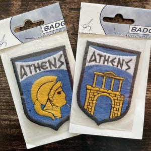 Può includere: Due toppe ricamate a forma di scudo in confezioni di plastica trasparente. Una toppa presenta un elmo spartano giallo su sfondo blu con il testo ATHENS. L'altra mostra un antico arco giallo su sfondo blu con il testo ATHENS.