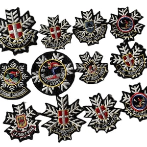 May include: A selection of embroidered patches with diverse designs. The patches have black and white snowflake-shaped borders, with colourful emblems and text. Some patches show shields with crosses, others depict skiers.