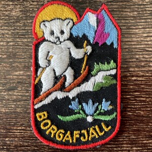 May include: An embroidered patch featuring a white bear skiing on a snowy slope against a black background. The design includes a yellow sun, blue and purple mountains, green foliage, and blue flowers. The text BORGAFJÄLL is stitched in yellow.