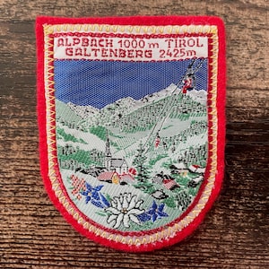Könnte beinhalten: Gestickter Aufnäher mit rotem Rand und detaillierter Berglandschaft. Der Aufnäher zeigt einen blauen Himmel, grüne Berge, eine Kirche und einen Skilift. Der Text oben lautet "ALPACH 1000 m TIROL GALTENBERG 2425m".