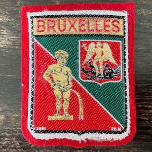 以下が含まれることがあります： 赤い縁取りの長方形の刺繍ワッペンで、金色の文字でBRUXELLESと書かれています。デザインには、赤い背景に金色の小便小僧のフィギュア、緑の背景に金色の天使のフィギュアが描かれた小さな盾が含まれています。