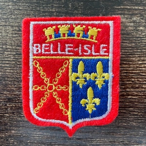 May include: A rectangular embroidered patch with a red border featuring the text BELLE-ISLE in white. The shield design includes a yellow crown, a yellow chain cross on a red background, and three yellow fleur-de-lis on a blue background.