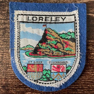Peut inclure: Un écusson en feutrine bleue avec une broderie représentant le texte LORELEY, ST. GOAR et ST. GOARSHAUS. Le motif illustre un paysage de falaise rocheuse, un petit drapeau et deux écussons héraldiques en bas aux accents rouges et bleus.