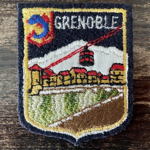 May include: A shield-shaped embroidered patch featuring the text GRENOBLE in white. The design depicts a red cable car, a white snow-capped mountain, a row of buildings with red roofs, and a green and brown landscape, all bordered in yellow.