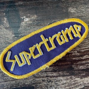 Può includere: Toppa ricamata con la scritta "Supertramp" in bianco, bordata di giallo, su sfondo blu scuro. La toppa ha un bordo giallo e una forma ovale. È su una superficie di legno.