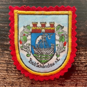 May include: A rectangular fabric patch with a red scalloped felt border and a yellow embroidered edge. The centre features a shield with a red castle, a blue field with a white sailing ship, and green waves. The text Bad Schandau a.E. is printed below.