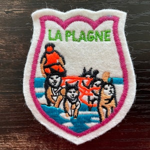 Può includere: Toppa ricamata a forma di scudo, raffigurante una squadra di cani da slitta che traina una slitta con una persona in abiti arancioni. La toppa ha il testo "LA PLAGNE" in verde, con un bordo rosa e bianco.