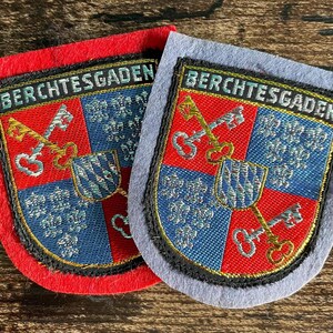 Könnte beinhalten: Zwei gestickte Aufnäher mit dem Wappen von Berchtesgaden. Jeder schildförmige Aufnäher zeigt den Schriftzug BERCHTESGADEN, zwei gekreuzte Schlüssel und einen kleinen Schild mit blau-weißen Rauten auf einem rot-blauen Hintergrund.