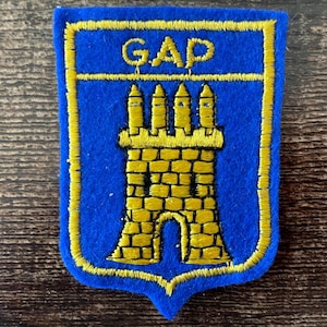 Peut inclure: Un écusson en feutrine bleue avec une bordure brodée jaune et un motif de château brodé en jaune. Le mot GAP est cousu en fil jaune en haut de l'écusson en forme de blason, présentant une surface texturée et des bords bien définis.