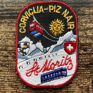 Op de afbeelding: Een geborduurde patch met een zwarte achtergrond en rode rand met de tekst CORVIGLIA-PIZ NAIR en St. Moritz. Het ontwerp bevat een rode kabelbaan, een gele zon, besneeuwde bergen, een wapenschild en een blauwe trein.