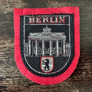 Op de afbeelding: Een rood en zwart Berlijn-patch met de Brandenburger Tor en het woord "BERLIJN" in witte letters. De patch heeft een zwarte rand met een decoratieve steek.