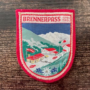 Könnte beinhalten: Gestickter Aufnäher mit rotem Rand, der eine Berglandschaft mit Gebäuden und dem Text "BRENNERPASS TIROL 1370m" zeigt. Der Aufnäher hat einen blauen Himmel, grüne Hügel und schneebedeckte Berge.