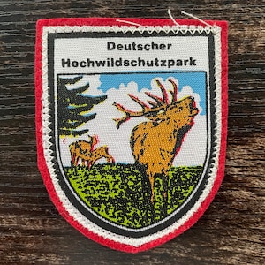 Puede incluir: Parche bordado con forma de escudo, con un ciervo con grandes cuernos en un prado. El parche tiene un borde rojo, costuras blancas y el texto "Deutscher Hochwildschutzpark".