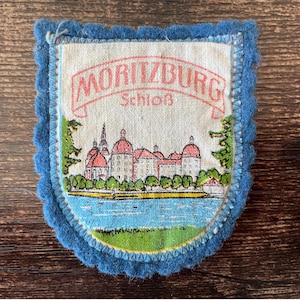 May include: A vintage souvenir patch featuring the text "MORITZBURG Schloss" in a banner. The patch depicts a castle with red roofs and a lake scene, framed by a blue, scalloped edge. The background is a dark wood surface.