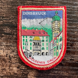 Könnte beinhalten: Gestickter Aufnäher mit einer farbenfrohen Darstellung von Innsbruck, Österreich. Der Aufnäher trägt oben den Text "INNSBRUCK" und unten "GOLDENES DACHL". Das Design zeigt Gebäude, einen Glockenturm und eine Bergkulisse, alles eingerahmt von einem roten Rand.