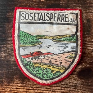 May include: Embroidered patch with a shield shape, featuring a landscape scene with a lake, trees, and a building. The patch has a red border and the text "SOSETALSPERRE HARZ".