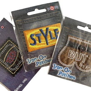 Può includere: Tre toppe termoadesive in confezione trasparente. Una toppa ha la scritta "STYLE" in blu su sfondo giallo. Un'altra toppa dice "BUT WEAR" in bianco su sfondo marrone. La terza toppa ha la scritta "TREND" in rosso.