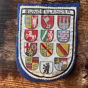 Peut inclure: Un écusson en tissu rectangulaire avec une bordure en feutre bleu portant le texte BUNDESLANDER en haut. Le centre blanc présente douze écussons héraldiques brodés représentant des États allemands, chacun avec des couleurs et symboles distincts.