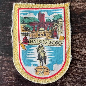 Puede incluir: Un parche de tela rectangular con un borde cosido amarillo y un marco interior rojo. El diseño presenta un castillo rojo, un cielo azul y una estatua ecuestre. Una cinta amarilla en el centro muestra el texto HÄLSINGBORG.