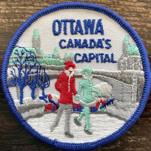 May include: A circular embroidered patch with a blue border featuring the text OTTAWA CANADA'S CAPITAL in blue. The design depicts two figures skating on ice in front of a bridge and buildings, rendered in red, light green, grey, and white thread.
