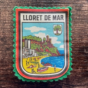 Könnte beinhalten: Ein rechteckiger Aufnäher mit dem Text "LLORET DE MAR" über einer farbenfrohen Illustration einer Küstenstadt. Das Bild zeigt einen Strand, Gebäude und eine Burg. Der Aufnäher hat einen grünen, gezackten Rand.