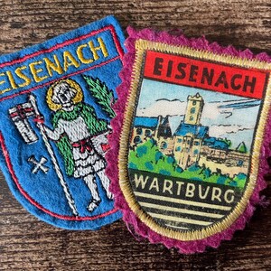 Könnte beinhalten: Zwei bestickte Stoffaufnäher. Der linke Aufnäher ist blau mit dem Text EISENACH und einer Figur mit Stab. Der rechte Aufnäher hat einen gezackten magentafarbenen Rand, einen roten Balken mit dem Text EISENACH und eine Darstellung der Wartburg.