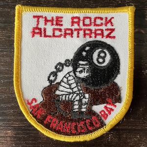 Könnte beinhalten: Ein schildförmiger gestickter Aufnäher mit gelbem Rand und weißem Hintergrund. Er zeigt den roten Text THE ROCK ALCATRAZ oben und SAN FRANCISCO BAY unten. Eine Figur in gestreifter Kleidung sitzt mit einem Kopf in Form einer schwarzen Acht-Kugel.