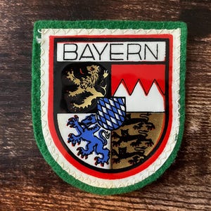 May include: A shield-shaped patch with a green felt border and a white stitched edge. The center features the word BAYERN above a coat of arms divided into four quadrants, displaying a gold lion, red and white peaks, a blue lion, and three black lions.