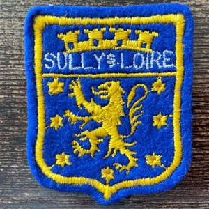 Può includere: Una toppa ricamata a forma di scudo con sfondo blu e bordo giallo. Presenta un leone giallo al centro, circondato da stelle gialle, con una corona gialla sopra la scritta SULLY-SUR-LOIRE in lettere maiuscole bianche.