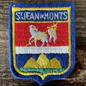May include: A rectangular embroidered patch with a blue border featuring the text S. JEAN DE MONTS in white. The design includes a red field with a white lamb holding a gold staff, a white horizontal stripe, and three yellow hills over green.