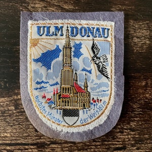 Op de afbeelding: Geborduurde patch met de Ulm Minster, een kathedraal in Ulm, Duitsland. De patch heeft een grijze rand en bevat de tekst "ULM DONAU" en "Höchstes Münster der Welt 161 m". Het ontwerp bevat blauwe, gouden en rode kleuren.