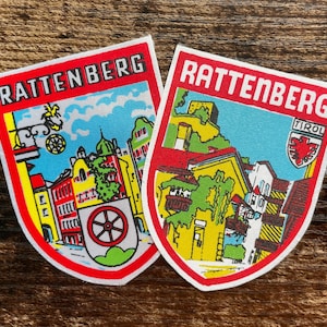 May include: Two shield-shaped embroidered patches featuring the text RATTENBERG in bold letters. The designs depict colorful town scenes with yellow buildings, green foliage, and red accents, including a wheel emblem and a small shield labeled TIROL.