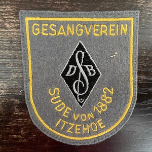 May include: A gray felt patch with yellow embroidered trim and text. The patch reads "GESANGVEREIN SUDE VON 1882 ITZEHOE" in yellow. A black and white diamond shape with musical notes and the letters "DB" is in the center.
