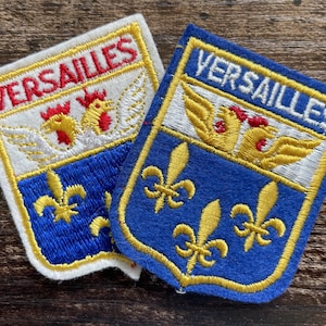 May include: Two embroidered patches featuring the text VERSAILLES. One patch has a white background with red text and a blue lower section, while the other has a blue background with white text. Both display golden winged roosters and fleur-de-lis.