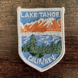 Könnte beinhalten: Gestickter Aufnäher mit einer Ansicht des Lake Tahoe. Der Aufnäher hat einen hellblauen Hintergrund mit den Worten "LAKE TAHOE" oben und "CALIF/NEV." unten. Das Design umfasst orangefarbene Berge, weißen Schnee, grüne Bäume und einen blauen See.