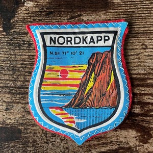 May include: A shield-shaped patch with a red border and a blue and white stitched edge. The patch features a landscape scene with a mountain, sea, and sunset, and the word "NORDKAPP" in a white box. The text "N.br. 71° 10' 21" is also present.