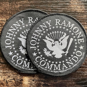 May include: Two black and white circular patches. The patches feature the text "JOHNNY RAMONE COMMANDO" around a central design of an eagle holding a baseball bat. The patches are on a wooden surface.