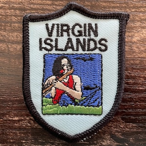 May include: A shield-shaped embroidered patch with a light blue background and a black border. The text VIRGIN ISLANDS is stitched in black at the top. Below, a figure plays a flute against a blue sky and green grass, rendered in thread.