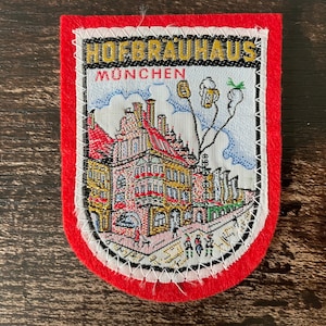 Può includere: Una toppa ricamata rossa, bianca e blu con il testo "HOFBRAUHAUS MÜNCHEN". La toppa presenta un'illustrazione dettagliata di un edificio con un tetto rosso e un cielo blu.