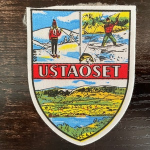 May include: A shield-shaped patch with the word "USTAOSET" in red. The top section depicts skiers and a fisherman. The bottom section shows a landscape with a lake and mountains. The patch is bordered in white.