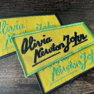 May include: Yellow embroidered patches with the name "Olivia Newton John" in black and green thread. The patches have a black border and a green border. The patches are rectangular.