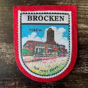 Könnte beinhalten: Ein roter Aufnäher mit weißem Rand, der das Wort "BROCKEN" und eine Illustration eines Gebäudes und Turms zeigt. Der Aufnäher enthält den Text "1142 m" und "Ich war oben". Der Aufnäher ist mit weißen Nähten eingefasst.