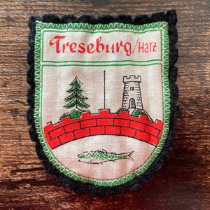 May include: A vintage patch featuring the text "Treseburg/Harz" in red, above a red brick bridge, a green fir tree, and a stone tower. The patch has a black scalloped border and a green border around the central design.