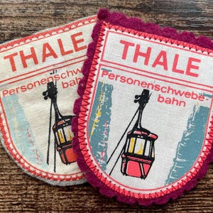 Può includere: Due toppe in tessuto a forma di scudo con la grafica di una funivia rossa su un cavo. Il testo recita THALE Personenschwebebahn. Una toppa ha un bordo color crema chiaro, l'altra un bordo in feltro smerlato magenta scuro con cuciture.