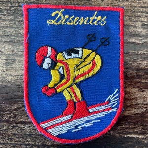 May include: Embroidered patch with a blue background and a red border. The patch features a skier in a yellow and red ski suit, with the word "Disentes" in gold script at the top. The skier is skiing down a slope with white lines.